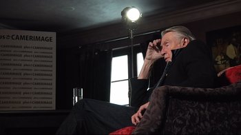 Movie still from “Side by Side” (2012), directed by Christopher Kenneally – An older man sitting in a chair talking on a phone; Medium shot, Over the shoulder angle