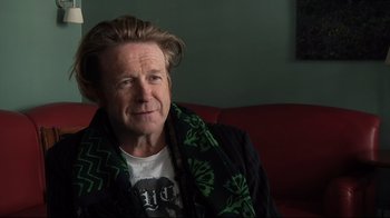 Movie still from “Side by Side” (2012), directed by Christopher Kenneally – A man sitting on top of a red couch wearing a black and green scarf; Close Up shot, Low angle