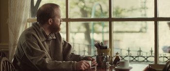 Movie still from “Sightseers” (2012), directed by Ben Wheatley – A man sitting at a table with a cup of coffee; Medium shot, Over the shoulder angle