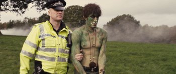 Movie still from “Sightseers” (2012), directed by Ben Wheatley – A man with green body paint and a police officer standing next to him in a grassy area; Medium shot, Over the shoulder angle