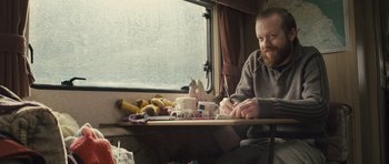 Movie still from “Sightseers” (2012), directed by Ben Wheatley – A man sitting in front of a window writing on a piece of paper; Medium shot, Low angle