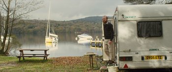 Movie still from “Sightseers” (2012), directed by Ben Wheatley – A man standing in front of a motorhome on the shore of a body of water; Wide shot, Low angle