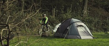 Movie still from “Sightseers” (2012), directed by Ben Wheatley – A man riding a bike next to a tent in the woods; Wide shot, Low angle