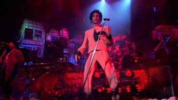 Movie still from “Sign 'o' the Times” (1987), directed by Albert Magnoli – A man holding a microphone on a stage; Wide shot, Low angle