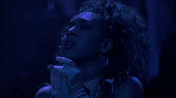 Movie still from “Sign 'o' the Times” (1987), directed by Albert Magnoli – A woman with curly hair is standing in a dark room; Close Up shot, Low angle