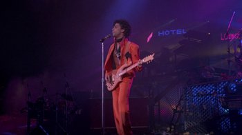 Movie still from “Sign 'o' the Times” (1987), directed by Albert Magnoli – A man in a red suit holding a microphone and a guitar; Wide shot, Low angle