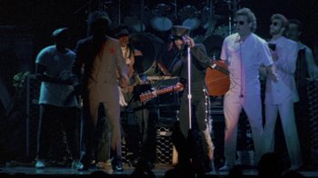 Movie still from “Sign 'o' the Times” (1987), directed by Albert Magnoli – A group of people on a stage with a microphone and a guitar; Wide shot, Low angle