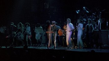 Movie still from “Sign 'o' the Times” (1987), directed by Albert Magnoli – A group of people standing on a stage with musical instruments; Wide shot, Low angle