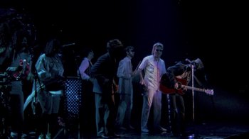 Movie still from “Sign 'o' the Times” (1987), directed by Albert Magnoli – A group of people standing on a stage with a guitar; Wide shot, High angle