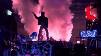Movie still from “Sign 'o' the Times” (1987), directed by Albert Magnoli – A man standing on top of a stage holding a gun; Wide shot, Low angle