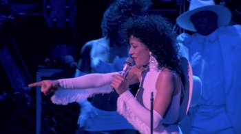 Movie still from “Sign 'o' the Times” (1987), directed by Albert Magnoli – A woman in a white dress holding a microphone; Medium shot, High angle