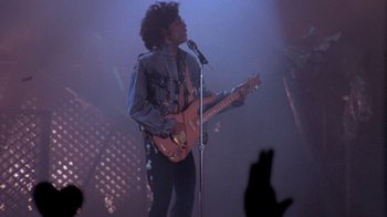Movie still from “Sign 'o' the Times” (1987), directed by Albert Magnoli – A man holding a microphone while playing a guitar on a stage; Medium shot, Low angle