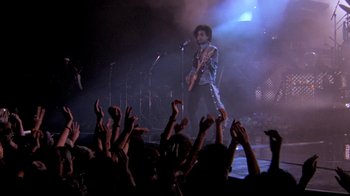 Movie still from “Sign 'o' the Times” (1987), directed by Albert Magnoli – A man with a guitar on a stage with a crowd; Wide shot, Low angle