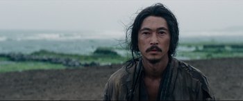Movie still from “Silence” (2016), directed by Martin Scorsese – A man with long black hair standing on a beach; Close Up shot, Over the shoulder angle