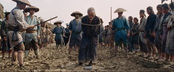 Movie still from “Silence” (2016), directed by Martin Scorsese – An old man is tied to a rope while a group of people are in the background; Wide shot, Low angle