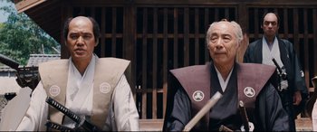 Movie still from “Silence” (2016), directed by Martin Scorsese – Two men sitting next to each other in front of a wooden fence; Medium shot, Low angle