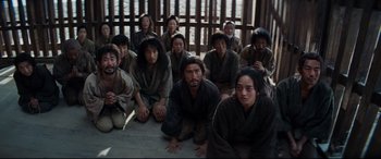 Movie still from “Silence” (2016), directed by Martin Scorsese – A group of people sitting on the ground in front of a wooden fence; Wide shot, High angle