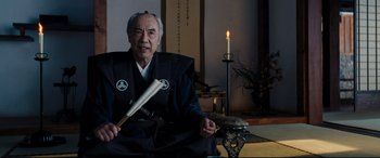 Movie still from “Silence” (2016), directed by Martin Scorsese – An older man sitting in a chair holding a baseball bat; Medium shot, Over the shoulder angle