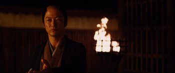 Movie still from “Silence” (2016), directed by Martin Scorsese – A man standing in front of a fire in a room; Medium shot, Low angle
