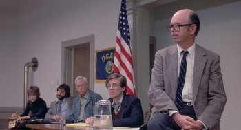 Movie still from “Silkwood” (1983), directed by Mike Nichols – A group of people sitting at a table in front of an american flag; Medium shot, Low angle