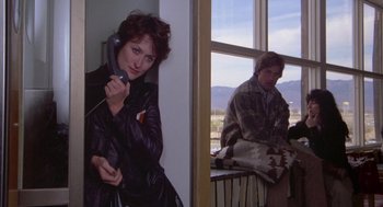 Movie still from “Silkwood” (1983), directed by Mike Nichols – A woman is talking on a phone while a man sits on a window ledge; Medium shot, Over the shoulder angle