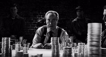 Movie still from “Sin City: A Dame to Kill For” (2014), directed by Frank Miller – A man sitting in front of stacks of coins; Medium shot, Over the shoulder angle