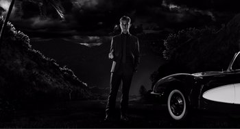 Movie still from “Sin City: A Dame to Kill For” (2014), directed by Frank Miller – A black and white photo of a man standing next to an old car; Wide shot, Low angle