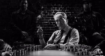 Movie still from “Sin City: A Dame to Kill For” (2014), directed by Frank Miller – An older man sitting at a table smoking a cigarette; Medium shot, Low angle