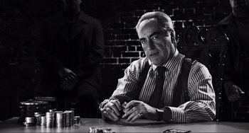 Movie still from “Sin City: A Dame to Kill For” (2014), directed by Frank Miller – A man sitting at a table in front of a brick wall; Medium shot, Over the shoulder angle