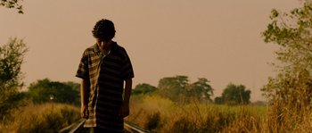 Movie still from “Sin Nombre” (2009), directed by Cary Joji Fukunaga – A young man standing in a field with trees in the background; Medium shot, Low angle