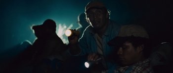Movie still from “Sin Nombre” (2009), directed by Cary Joji Fukunaga – A man holding a flashlight while sitting in a crowd of onlookers; Close Up shot, Low angle