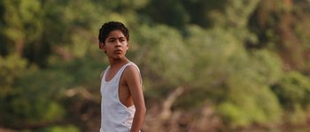 Movie still from “Sin Nombre” (2009), directed by Cary Joji Fukunaga – A young boy in a white tank top stands in front of trees; Medium shot, Over the shoulder angle