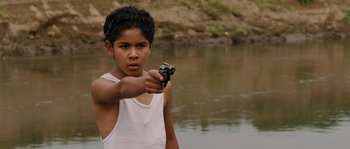 Movie still from “Sin Nombre” (2009), directed by Cary Joji Fukunaga – A young man holding a gun in front of a body of water; Close Up shot, High angle