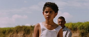 Movie still from “Sin Nombre” (2009), directed by Cary Joji Fukunaga – A young boy in a white tank top looking at the camera; Close Up shot, Over the shoulder angle