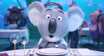 Movie still from “Sing” (2016), directed by Christophe Lourdelet – A koala sitting at a table with a plate in front of him; Medium shot, Over the shoulder angle
