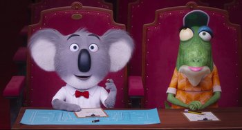 Movie still from “Sing” (2016), directed by Christophe Lourdelet – A koala sitting at a table with a pen in his mouth; Medium shot, Low angle