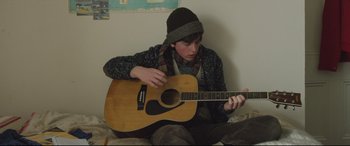 Movie still from “Sing Street” (2016), directed by John Carney – A person sitting on the ground playing a guitar; Medium shot, High angle