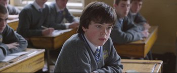 Movie still from “Sing Street” (2016), directed by John Carney – A boy sitting in front of a group of people; Close Up shot, High angle