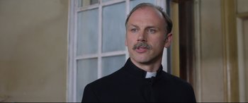Movie still from “Sing Street” (2016), directed by John Carney – A man in a priest's outfit looking at the camera; Close Up shot, Low angle