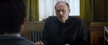 Movie still from “Sing Street” (2016), directed by John Carney – A man in a priest's outfit smoking a cigarette; Close Up shot, Over the shoulder angle