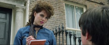 Movie still from “Sing Street” (2016), directed by John Carney – A young woman is holding a book and looking at the camera; Close Up shot, Over the shoulder angle