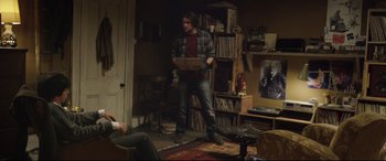 Movie still from “Sing Street” (2016), directed by John Carney – A man standing in a living room holding a box; Wide shot, Over the shoulder angle