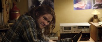 Movie still from “Sing Street” (2016), directed by John Carney – A man with long brown hair is smiling for the camera; Close Up shot, Over the shoulder angle