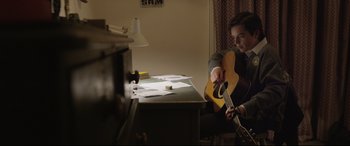 Movie still from “Sing Street” (2016), directed by John Carney – A man sitting at a desk playing a guitar; Medium shot, Low angle