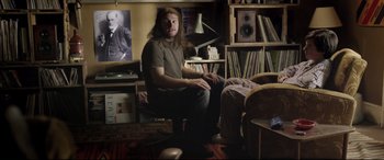 Movie still from “Sing Street” (2016), directed by John Carney – A man sitting on a chair in a living room; Medium shot, Low angle