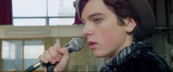 Movie still from “Sing Street” (2016), directed by John Carney – A person holding a microphone up to their mouth; Close Up shot, Over the shoulder angle
