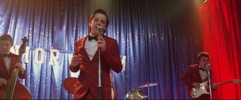 Movie still from “Sing Street” (2016), directed by John Carney – A man in a red suit is singing into a microphone; Medium shot, Low angle