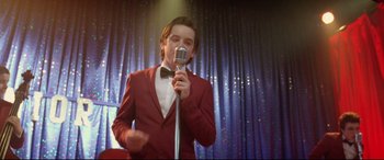 Movie still from “Sing Street” (2016), directed by John Carney – A man in a red suit is holding a microphone; Medium shot, Low angle