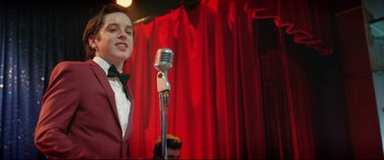Movie still from “Sing Street” (2016), directed by John Carney – A young man in a red suit is on a stage with a microphone; Medium shot, Low angle