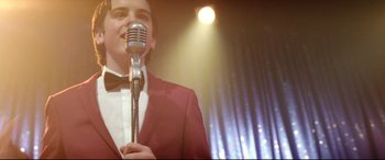 Movie still from “Sing Street” (2016), directed by John Carney – A man in a red jacket is holding a microphone; Medium shot, Low angle